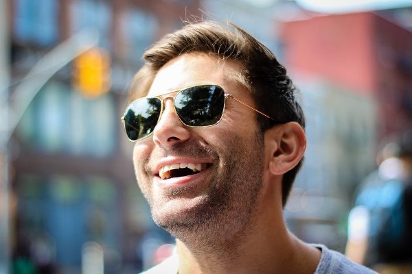 Hombre sonriente con lentes negros, fondo desenfocado.